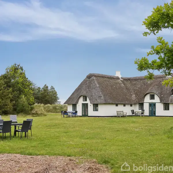 Et stråtækt hus står stille på en grøn græsplæne omgivet af træer og buske. I forgrunden er der et bord med stole, der skaber en afslappende atmosfære.