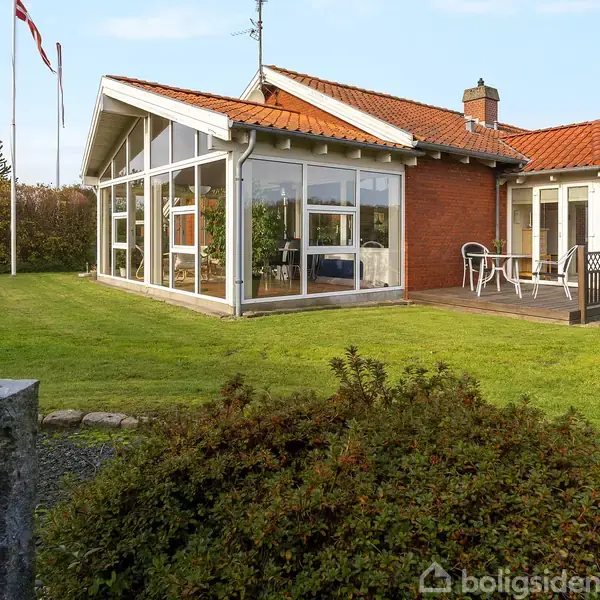 Et rødt murstenshus med store glasvinduer står i en grøn have. Flagstang med dansk flag i baggrunden, og terrassen har et bord og stole.