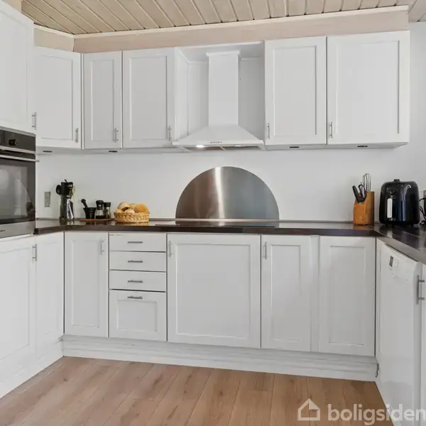 Hvidt køkken med indbyggede skabe og rustfri stålover. Kaffemaskine, brødrister og planter på bordpladen. Vindue til højre lukker lys ind.