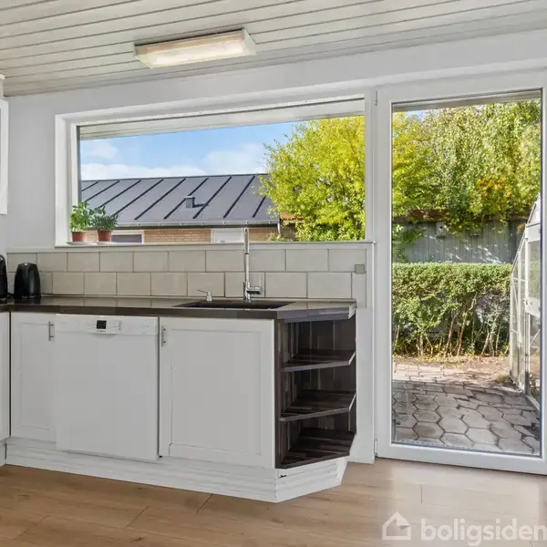 Køkken med hvide skabe står stille, udstyret med en vask og køkkenudstyr. Rummets store vindue og glasdør åbner ud til en flisebelagt terrasse og grøn have i baggrunden.