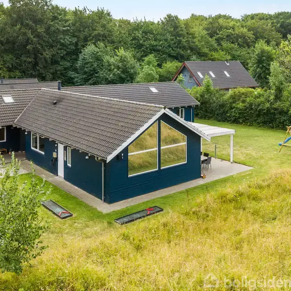 Blåt hus med store vinduer og mørkt tag står på grøn græsplæne. Terrasse dækker en del af siden. Skov og enkelte huse omkranser området. Legeplads med rutsjebane i baggrunden.