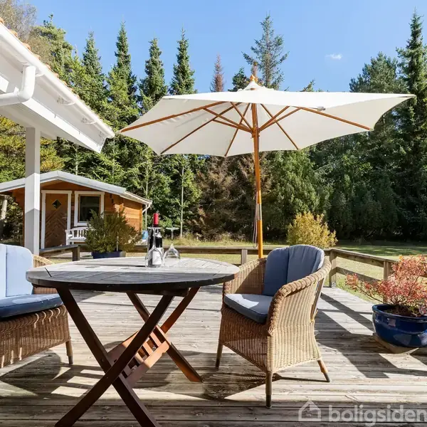 Terrasse med rundt træbord og to flettede stole med blå puder, står under en stor parasol. Området er omgivet af græsplæne og skov med fyrretræer i solskin.