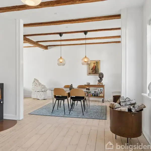 Brændeovn opvarmer et stilfuldt rum med træbjælker. To lamper hænger over et spisebord med stole. Et tæppe og kunstværker pryder rummet, skaber en hyggelig atmosfære.
