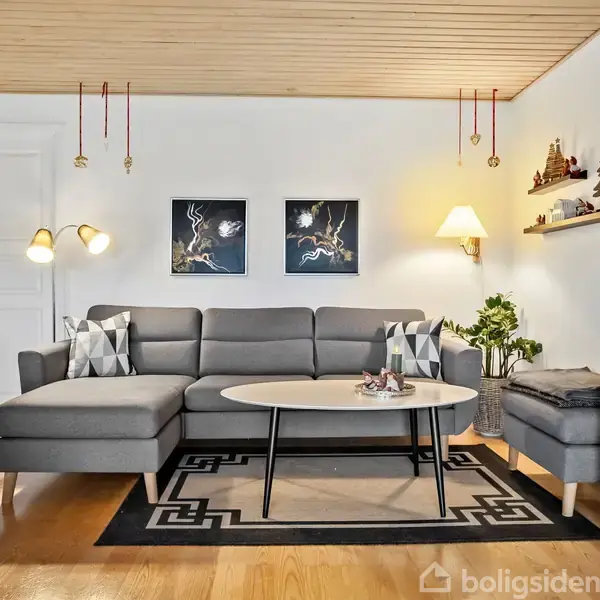 En grå hjørnesofa står mod en hvid væg, pyntet med to billeder. Sofaen har puder og er placeret på et tæppe i et hyggeligt indrettet rum med træloft.