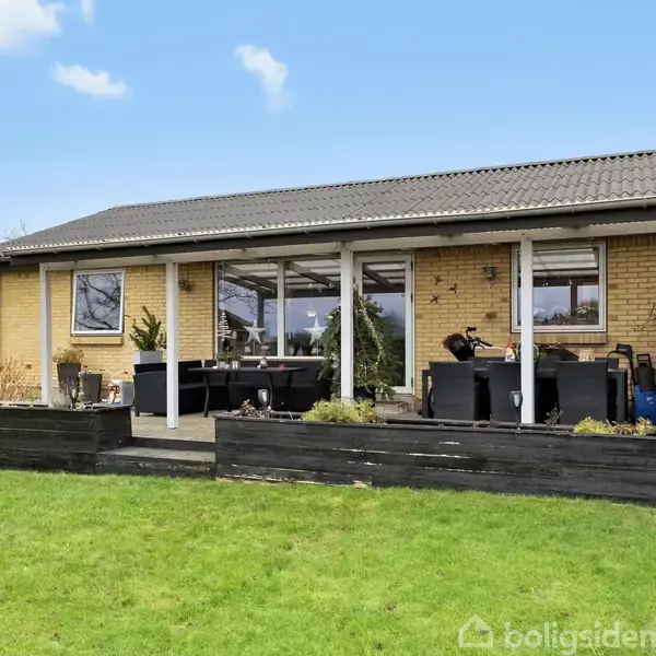 En enkeltstående gul murstensvilla med fladt tag og store vinduer. Foran huset står havemøbler på en terrasse. Græsplæne giver en åben, grøn ramme.