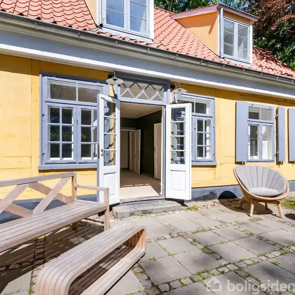 Gul bygning med åbne, hvide døre og blå skodder. Foran huset står to træbænke og en rund stol på en flisebelagt terrasse, omgivet af grønne træer.