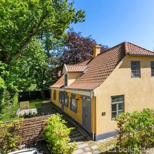 Et gult hus med rødt tegltag står omgivet af en frodig have. Sollys skinner gennem de grønne træer, der skaber en hyggelig stemning. Adresseplade: "34".