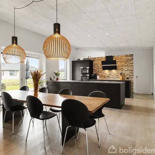 Lang spisebord med sorte stole står i et moderne køkken-alrum. To store, hængende lamper lyser over bordet. Murstensvæg i baggrunden, og lysindfald fra vinduer. Ingen tekst.