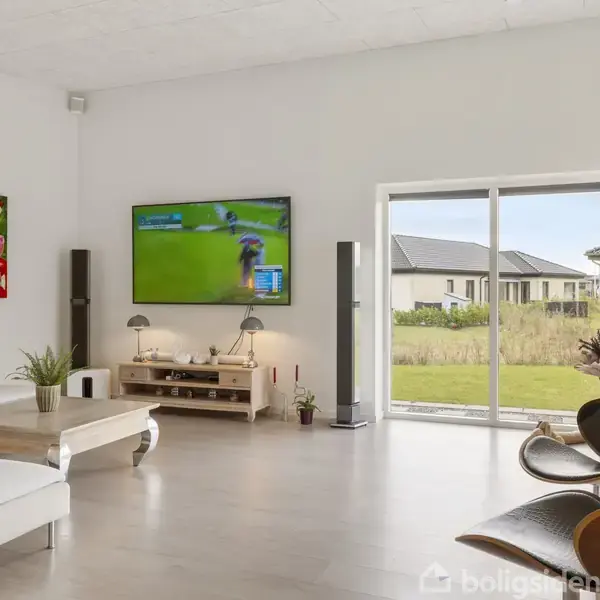 Stue med hvid sofa, tv viser golf, farverig kunst på væggen, store vinduer med kig til græsplæne og huse udenfor. En træbord og moderne stol er også til stede.