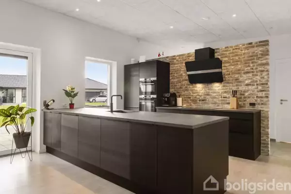 Et moderne køkken med mørke skabe og en stor køkkenø. Der er en murstensvæg bag komfuret. Rummet har store vinduer med udsigt til en terrasse udenfor.