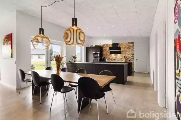 To pendler hænger over et træbord omgivet af sorte stole. I baggrunden ses et moderne køkken med murstensvæg og ø, alt i et lyst rum med store vinduer.