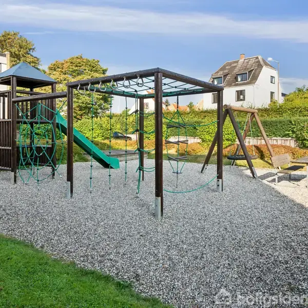 Legeplads med klatrestativ og rutsjebane i centrum. Gynger og sandkasse i baggrunden. Beliggende i et grønt område omgivet af huse og buske. Skilt: "Enhver færdsel på arealet sker på eget ansvar".