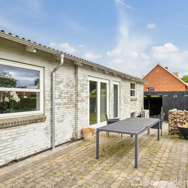 Dør åbnes ind i en lys entre, hvor en stol står ved væggen. Trætrappen fører op til et rum med vindue. Lyse vægge og trægulv skaber et moderne udtryk.