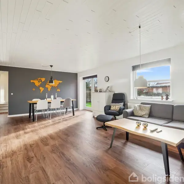 Sofagruppe står i stuen, badet i naturligt lys fra vinduerne, omgivet af lyst trægulv og et kort på væggen. Et spisebord med stole ses i baggrunden.