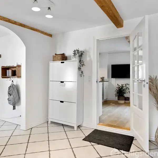 En lys entré med hvidt skoskab, hvor en dør fører til en stue med planter. Rummets gulv har lyse fliser, og der er synlige træbjælker i loftet.