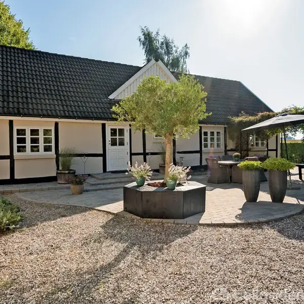 Hus med sort-hvid facade står stille i en solbeskinnet have, omgivet af grusvej og planter i krukker. En bænk og kysestol står til højre.