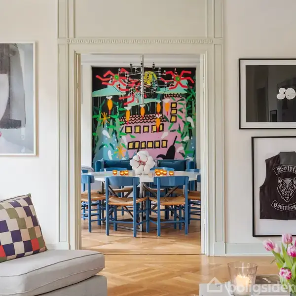 Spisebord med blå stole står i midten af en lys stue. En moderne blå sofa og farverig vægkunst ses til højre, omgivet af store vinduer og lyse træmøbler.