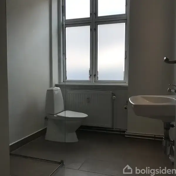 Toilet stående ved siden af en lille håndvask, placeret tæt på et stort vindue i et enkelt og lyst badeværelse. Der ses en radiator under vinduet.
