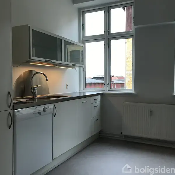 Et funktionelt køkken med hvide skabe og indbyggede apparater som opvaskemaskine. Over vasken er der belysning. Store vinduer slipper naturligt lys ind i rummet.