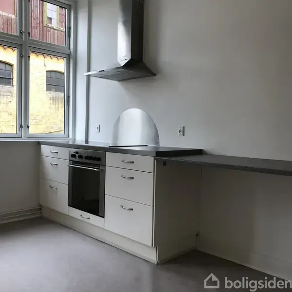 Køkken med hvide skabe og en indbygget ovn under en kogeplade. Emhætte er monteret over komfuret. Store vinduer giver udsigt til en gul murstensbygning udenfor.