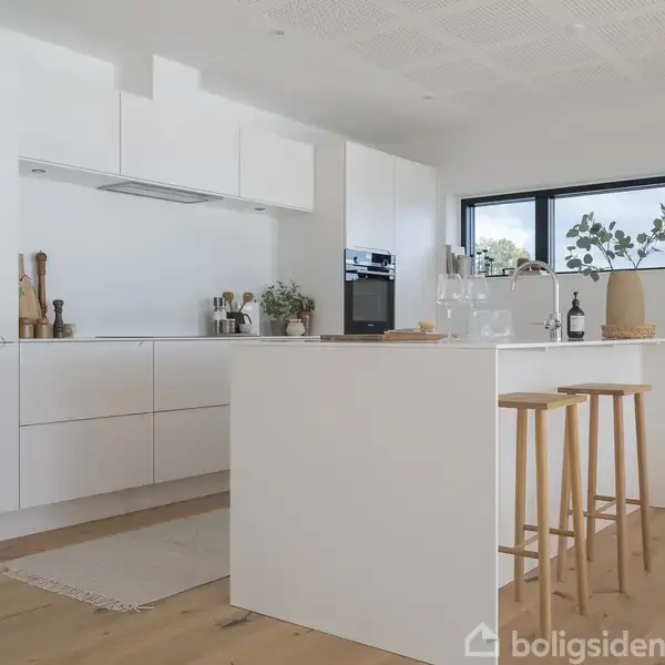 Hvidt køkken med indbygget ovn og kogeplade, omgivet af moderne skabe. Køkkenø med barstole og pynt. Stor vindue giver naturligt lys og udsigt til naturen udenfor.