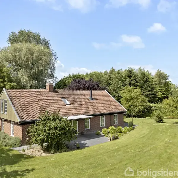 Et rødt murstenshus står stille omgivet af frodige træer og en velplejet græsplæne under en blå himmel.