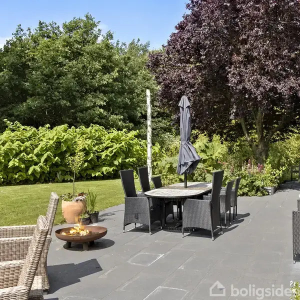 En terrasse med sorte møbler og parasol står på fliser, omgivet af grønne buske og træer. En ildskål er placeret ved siden af kurvestole.