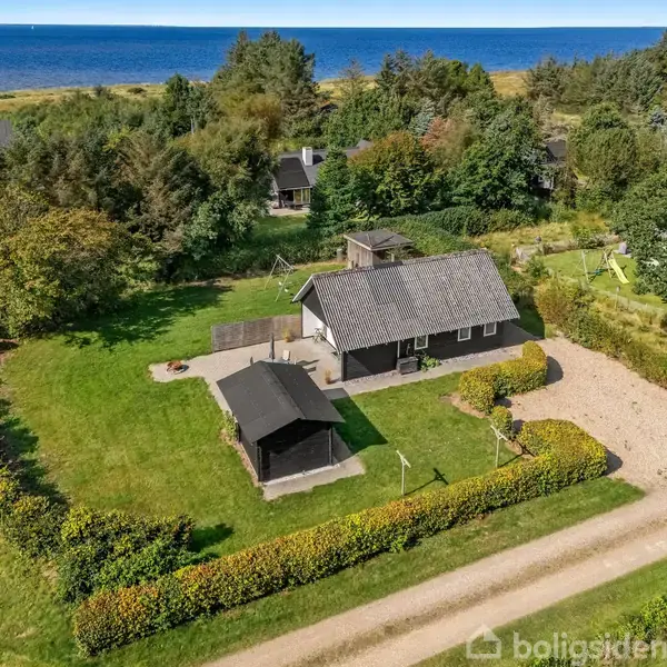 Et sort hus med gråt tag står i et grønt område omgivet af træer og buske. Græsplæne og lille skur ses, med havet i baggrunden. En grusvej løber forbi.