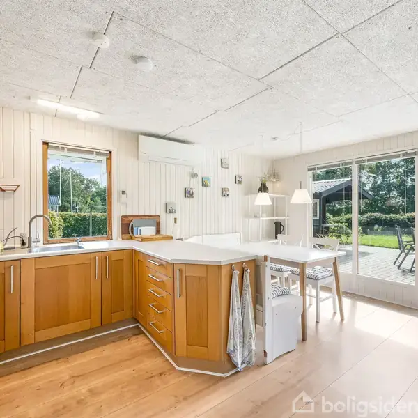 Et lyst køkken med træskabe er forbundet til en spiseplads ved store vinduer, der åbner ud til en terrasse med havemøbler. Udsigt til en grøn have.