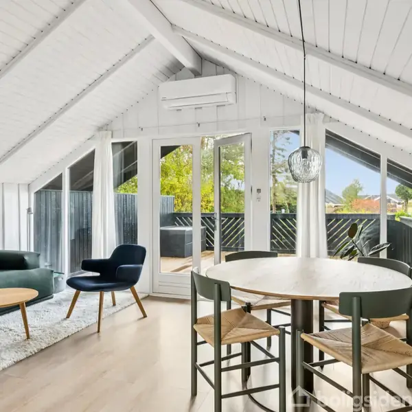 Stue med stejlt loft. Sofa og lænestol står ved et lavt bord på et hvidt tæppe. Rundt spisebord med tre stole. Store vinduer fører til terrasse med udsigt.