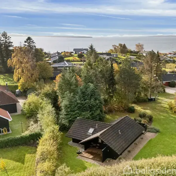 Sort hus med have omgivet af træer; beliggende i en grøn, kuperet landsby med udsigt til havet i baggrunden. Skiftende høstfarver præger landskabet.
