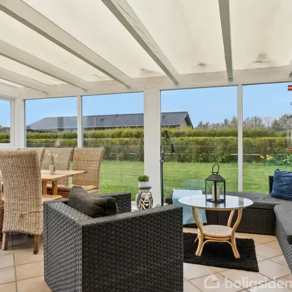 Fletstole og bord står i en lys udestue med glasvægge. En lanterne pryder et lille rundt bord. Udsigt til en velholdt have med græsplæne og granitfigur.