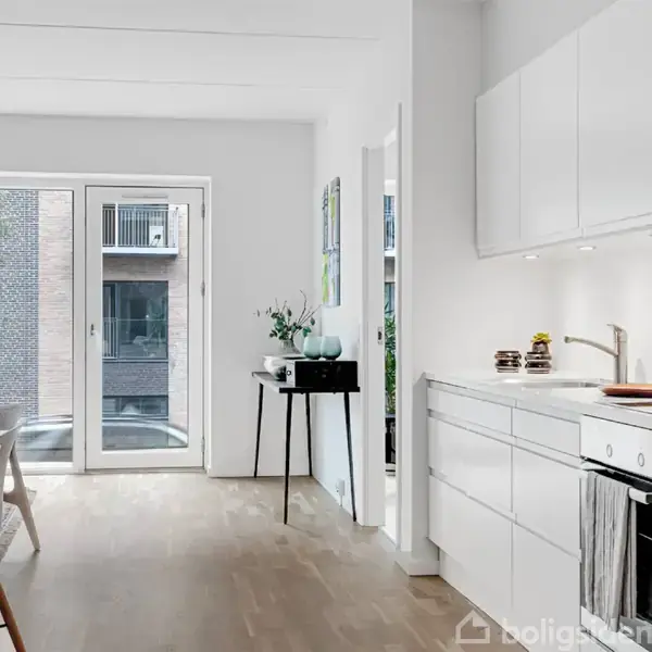 Moderne køkken-alrum med hvide skabe og træbord, oplyst af store vinduer med udsigt til en bygning. Plante og skåle står på bordet. Trægulve gennem hele rummet.