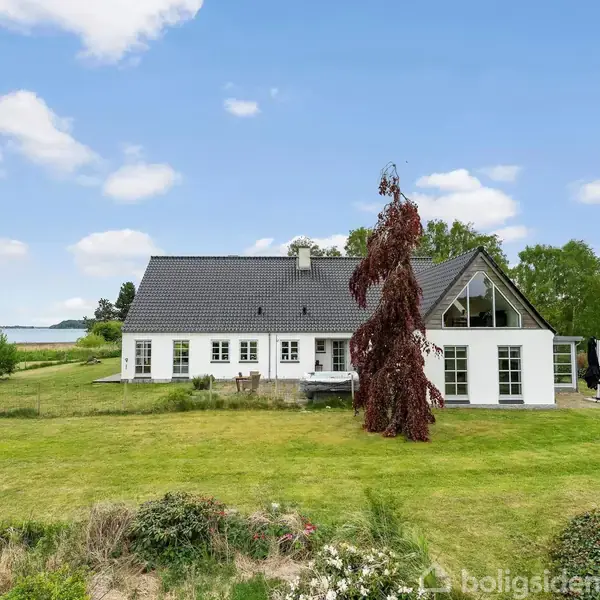 Et hvidt hus med gråt tag står på en grøn græsplæne. En rødbladet træ er foran huset. I baggrunden ses en sø og træer under en blå himmel med skyer.