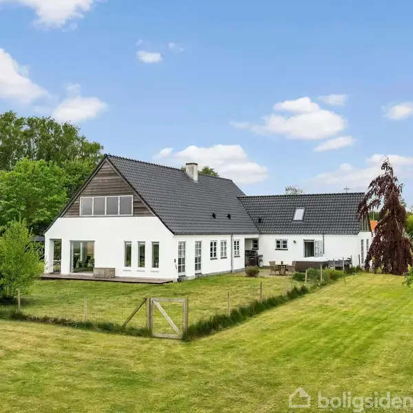 Et hvidt hus med sort tag står på en velplejet græsplæne omgivet af grønne træer og buske under en blå himmel med spredte skyer.