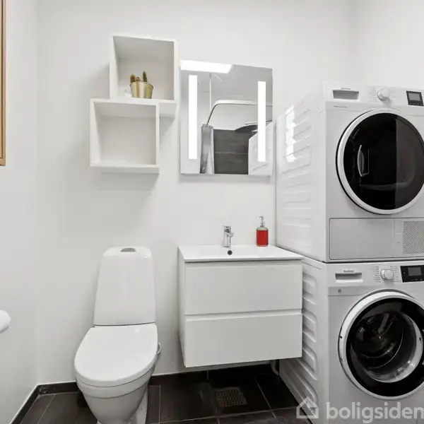 Toilet står ved siden af en vægmonteret håndvask under et spejl. En vaskemaskine og tørretumbler er stablet ved siden af. Hvide hylder på væggen. Rummet er lyst.