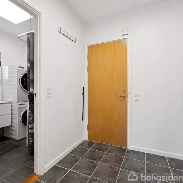 Dør fører ind til en entré med gråt flisegulv. Til venstre ses et badeværelse med vask og vaskesøjle. Højre side har et køleskab. Væggene er hvide.