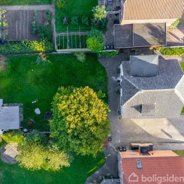 Et hus med en grå tag og en stor have set fra oven. Haven indeholder træer og forskellige sektioner med planter. Huset er omgivet af andre bygninger og en vej.