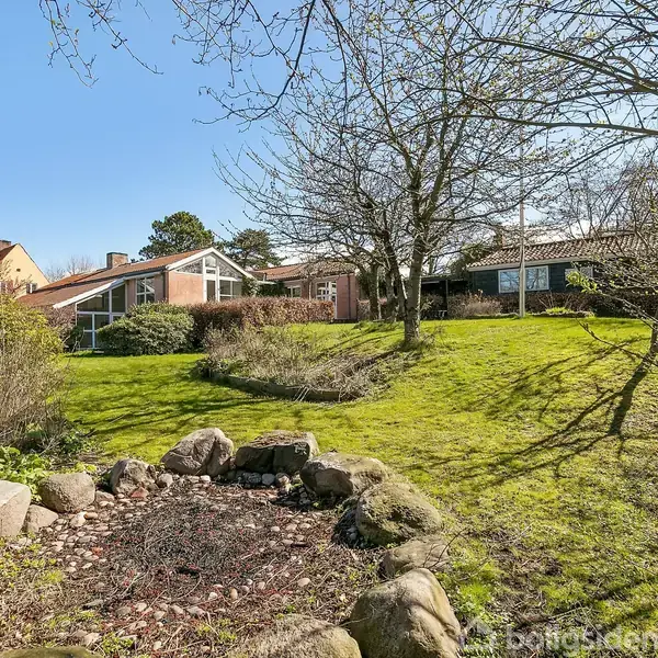 Hus i baggrunden omgivet af grøn have; et træ står foran huset. Foran er der stenarrangement omkring en lille grusbunke. Sollys oplyser scenen, hvilket skaber en forårsagtig atmosfære.