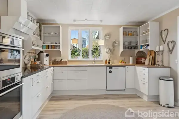 Et lyst køkken med hvide skabe og moderne apparater. Der er en ovn, opvaskemaskine og vindue med udsigt til planter udenfor. Hylderne indeholder tallerkener og krukker.