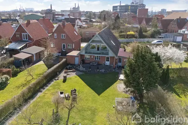 Et rødt murstenshus med skråt tag står i en have. Huset er omgivet af andre huse og træer. I baggrunden er der en by fyldt med bygninger.