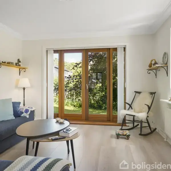 Stue med en blå sofa ved siden af et rundt sofabord. En gyngestol står ved et vindue. Store glasdøre fører ud til en have. Hylder med billeder og lampe på væggen.