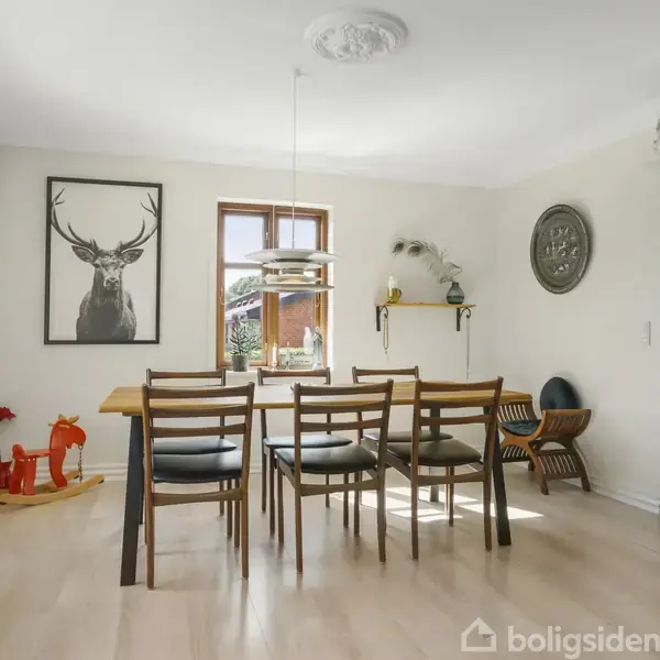 Spisebord med seks stole står i en lys spisestue. Billedet af en hjort hænger på væggen, omgivet af planter og dekorative elementer. Vinduerne giver naturligt lys.