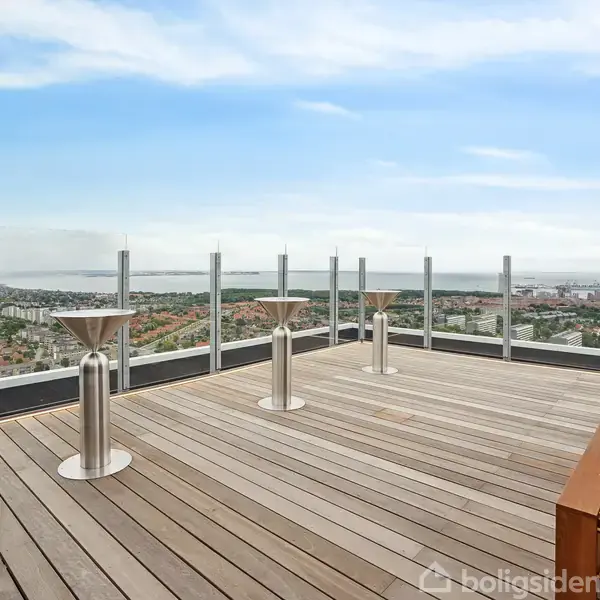 Tagterrasse i træ med udsigt over en by og kystlinje. Tre moderne stålsøjler langs glasrækværk. Klar himmel og få skyer i baggrunden.