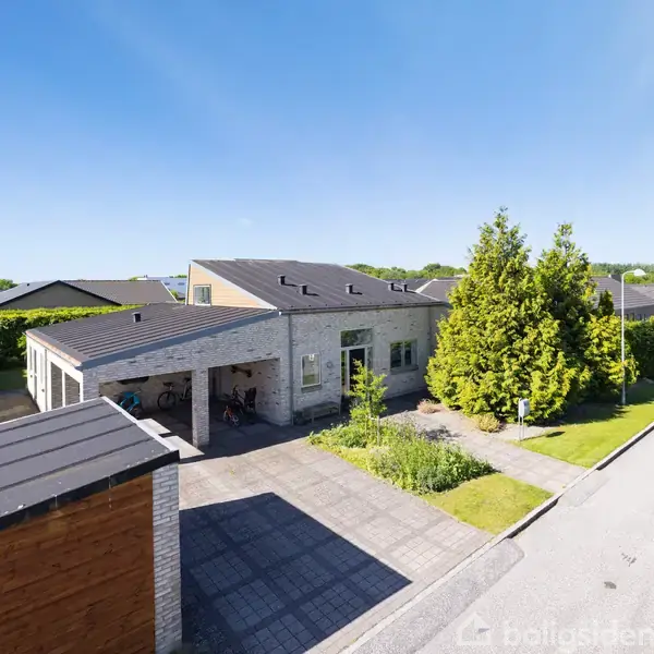 Et moderne hus med fladt tag står ved siden af en indkørsel og en garage. Grønt buskads og træer omgiver huset, som ligger i et roligt villakvarter.