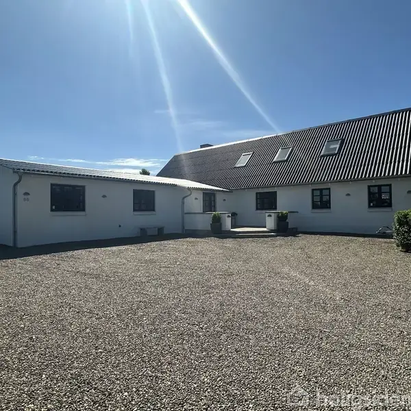 Hvid bygning med skråt tag og flere vinduer, beliggende på en grusbelagt gårdsplads. Sollys skinner ned fra en klar blå himmel, og der er grønt hegn i højre side.