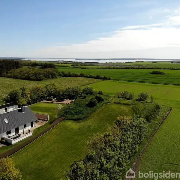 En hvid landejendom ligger fredeligt omgivet af grønne marker og træer, med en udsigt over en fjord i baggrunden under en klar himmel.