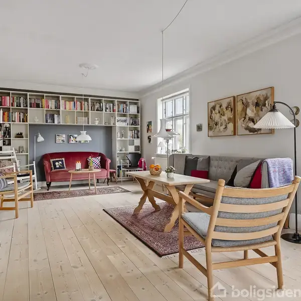 Stue med træmøbler og grå sofa, oplyst af store vinduer. Væghylde fyldt med bøger og dekorationer. Rød sofa i baggrunden, gulvtæppe og planter tilføjer farve og liv.