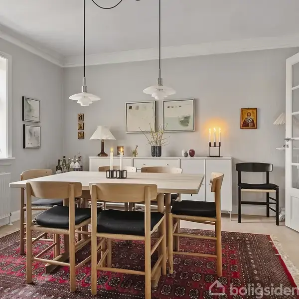 Spisebord med seks stole står på orientalsk tæppe i spisestue. På væggen hænger kunst og en tændt lampe. Vindue til venstre, reol til højre.