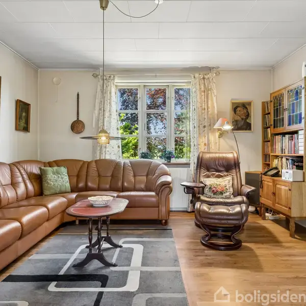 En brun lædersofa står i en stue med et rundt sofabord foran den. En lænestol med læselampe står ved siden af en bogreol. Store vinduer lader dagslys komme ind.